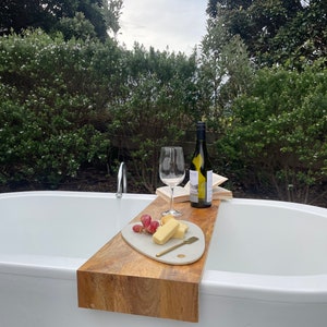 May include: A wooden bath tray with a white marble serving board, a glass of wine, a bottle of wine, and a plate of cheese and grapes. The tray is placed on the edge of a white bathtub.