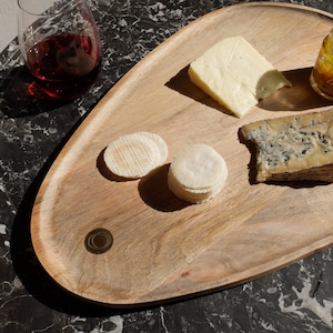 May include: A wooden serving tray with a rounded edge, featuring a variety of cheeses, crackers, and a small jar of preserves. The tray is made of light brown wood and has a natural finish.