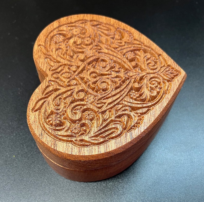 May include: A wooden heart-shaped box with intricate carved details. The box is made of a rich brown wood and has a smooth, polished finish.