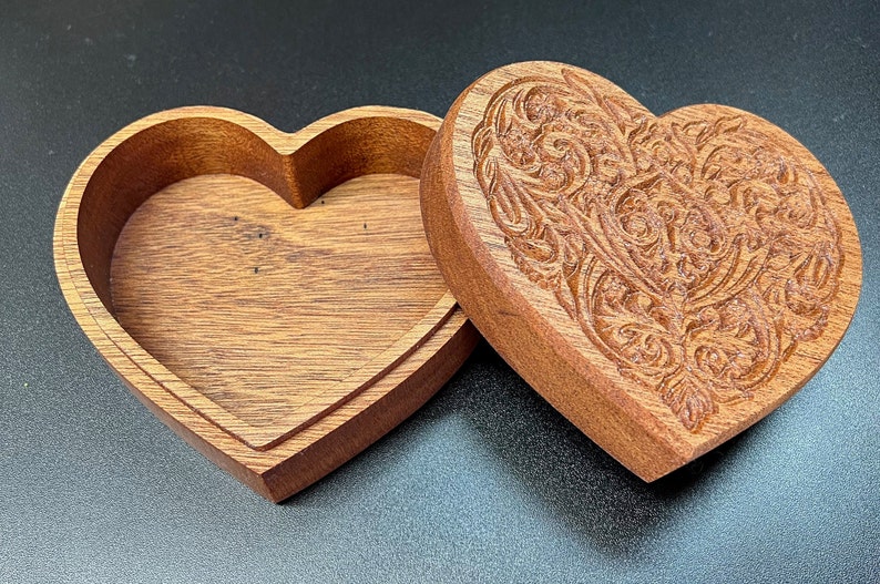 May include: A wooden heart-shaped box with an intricate carved design on the lid. The box is open and the inside is empty.