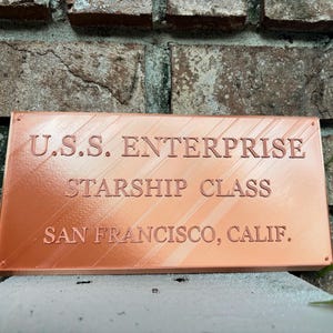 May include: A rectangular copper-colored plaque with raised lettering that reads U.S.S. ENTERPRISE, STARSHIP CLASS, and SAN FRANCISCO, CALIF. The surface features a diagonal texture pattern and small circular indentations at each corner.