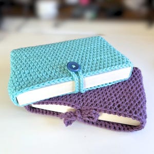 May include: Two rectangular book covers made of crocheted yarn, one in turquoise and one in purple. The turquoise cover features a single blue button closure with a matching yarn loop, while the purple cover has a knotted yarn tie closure.