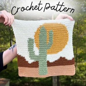 May include: A square crochet piece featuring a desert landscape design with a sage green cactus in the center, a large mustard yellow sun, and layered brown and tan hills against an off-white background. The text Crochet pattern is visible.