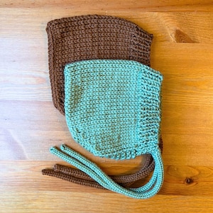 Puede incluir: Dos gorros de bebé tejidos. Uno es de color marrón liso y el otro es de color verde azulado claro. El gorro verde azulado tiene lazos largos. Los gorros están hechos de un hilo suave y texturizado y probablemente están hechos a mano.