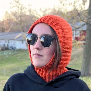 May include: Close-up of an orange crochet balaclava. The balaclava is thick and textured, with a ribbed design. It covers the head and neck, leaving the face partially visible.  The balaclava is shown on a person wearing sunglasses. The balaclava is a warm winter accessory, perfect for outdoor activities. Crochet balaclava hood pattern in burnt orange. Easy beginner-friendly crochet pattern with ribbed neck.