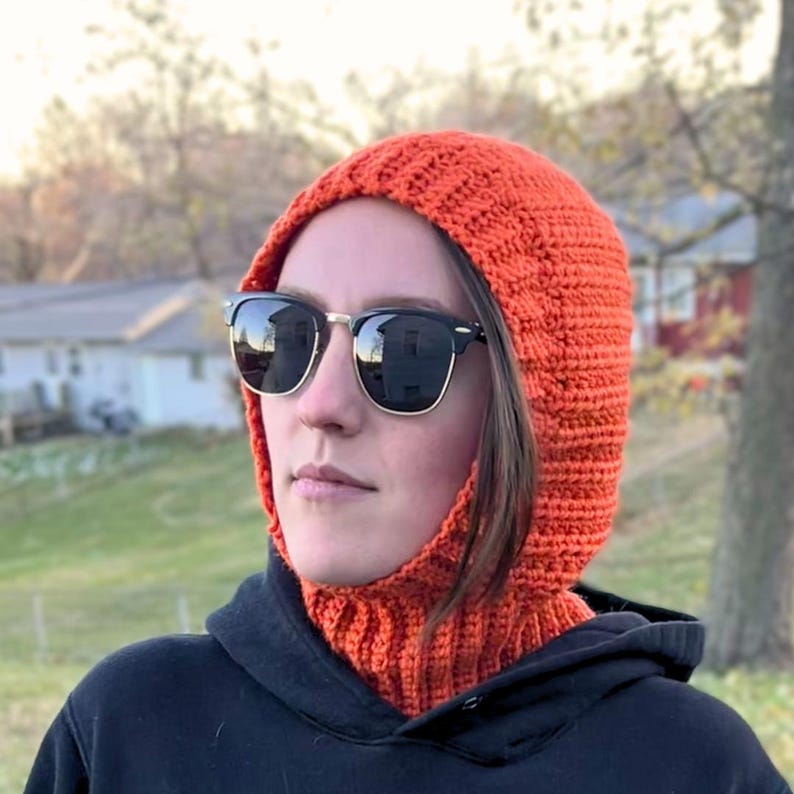 May include: Close-up of an orange crochet balaclava. The balaclava is thick and textured, with a ribbed design. It covers the head and neck, leaving the face partially visible.  The balaclava is shown on a person wearing sunglasses. The balaclava is a warm winter accessory, perfect for outdoor activities. Crochet balaclava hood pattern in burnt orange. Easy beginner-friendly crochet pattern with ribbed neck.