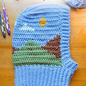 Op de afbeelding: Een lichtblauwe gehaakte bivakmuts met een landschapsontwerp van groene en bruine bergen, een witte wolk en een gele zon. De tekst Crochet pattern is bovenaan zichtbaar. Twee haaknaalden en een bol lichtblauw garen zijn aanwezig.