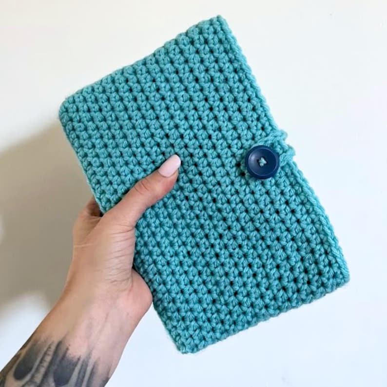 May include: A rectangular, turquoise crochet cover featuring a textured, repeating stitch pattern. The cover is secured with a single, dark blue circular button attached to a small loop on the right side, providing a functional closure for the item.