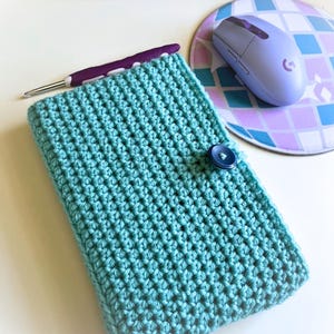 Op de afbeelding: Een turquoise gehaakte tas met een blauwe knoopsluiting. De tas is gemaakt met een textuur-steek en is perfect voor het opbergen van kleine items.