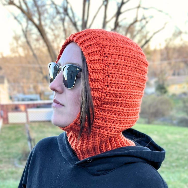 May include: Close-up of a crocheted balaclava. The balaclava is burnt orange and features a ribbed texture. It has a snug-fitting hood and a long neck covering. The balaclava is shown against a blurred outdoor background. Crochet balaclava hood pattern in burnt orange. Easy beginner-friendly crochet pattern with ribbed neck.