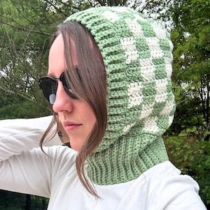 Puede incluir: Una capucha de crochet con un patrón de tablero de ajedrez verde y blanco. La capucha está hecha de un hilo suave y tiene un cuello acanalado y un patrón de cuadros en la capucha. La capucha está diseñada para usarse sobre la cabeza y el cuello.