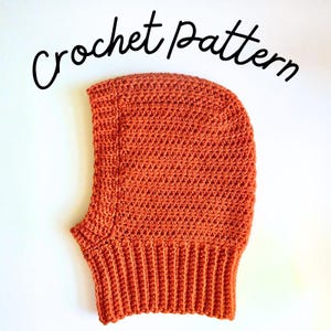 May include: A burnt orange crochet balaclava featuring a textured stitch pattern and a ribbed neck trim. The text "Crochet pattern" is written in black cursive script above the headwear, indicating this is a guide for creating the item.