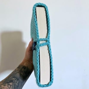 May include: A light blue crochet book sleeve featuring a textured yarn pattern and a dark blue button closure. The sleeve is wrapped around a thick book with visible white pages, showcasing the snug fit and the detailed hand-stitched border.