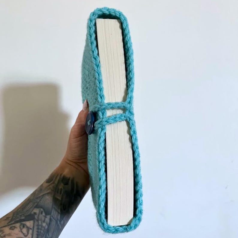 May include: A light blue crochet book sleeve featuring a textured yarn pattern and a dark blue button closure. The sleeve is wrapped around a thick book with visible white pages, showcasing the snug fit and the detailed hand-stitched border.