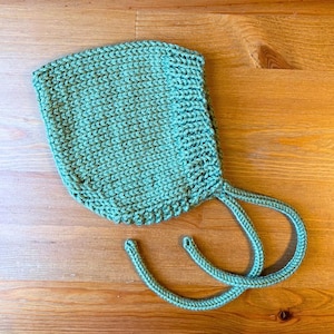 Può includere: Un cappellino per neonati lavorato a maglia in un morbido colore verde acqua tenue. Il cappellino presenta una trama a maglia fitta e lacci su entrambi i lati per una vestibilità sicura. L'articolo è esposto su una superficie di legno.