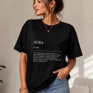 Peut inclure: T-shirt noir avec le mot "AURA" imprimé en blanc, ainsi qu'une définition. Le t-shirt est porté avec un jean bleu. La définition décrit l'énergie invisible que quelqu'un porte.