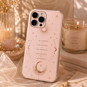 Peut inclure: Coque de téléphone transparente avec un fond rose ornée de motifs célestes et du texte "I AM ALIGNED, I AM FOCUSED, I AM ABUNDANT, I AM GRATEFUL, I AM ENOUGH." La coque a un motif de soleil et de lune doré.