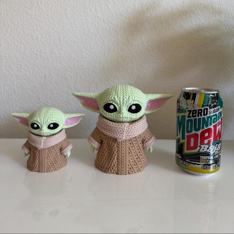 May include: Two green crocheted figures with large ears and black eyes, resembling a popular character, stand next to a can of Mountain Dew Baja Blast. The figures are wearing brown and pink clothing. The can is green, yellow, and blue.
