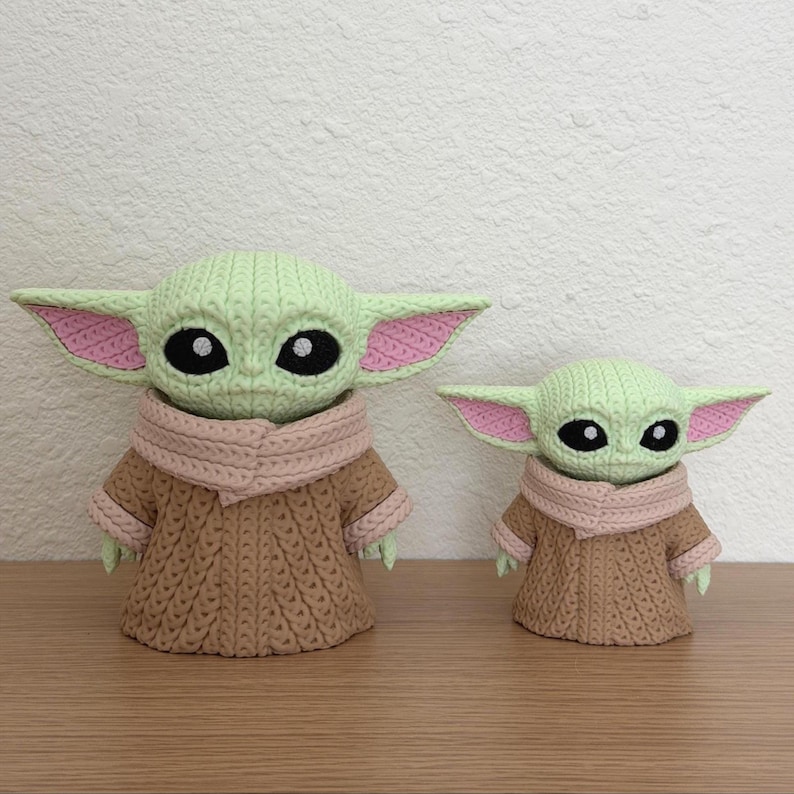 May include: Two crocheted figures of a character with large ears, large eyes, and a tan robe. The figures are in different sizes. The larger figure is approximately 15 cm tall, and the smaller figure is approximately 10 cm tall. The figures are on a wooden surface.
