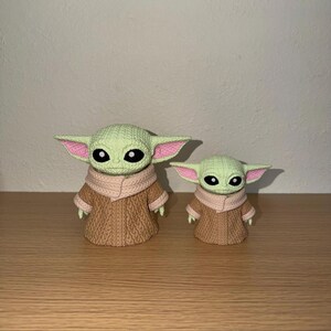 May include: Two green figurines of a character with large ears and black eyes, wearing a brown knitted coat and a light pink scarf. The figurines are of different sizes, with the larger one on the left.