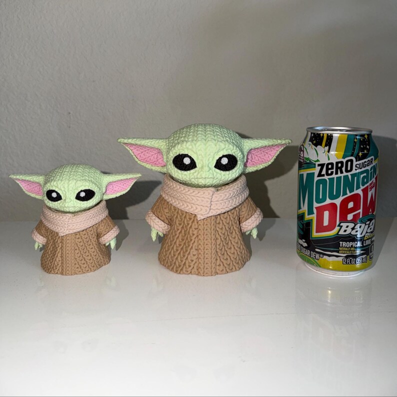 May include: Two crocheted figures of a character with large ears, green skin, and a tan robe. The figures are in different sizes. A can of Zero Sugar Mountain Dew Baja Tropical Lime soda is next to the figures.