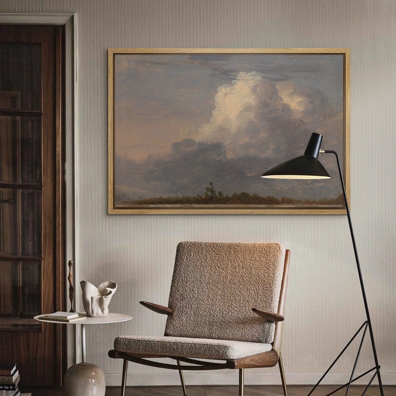 May include: A beige armchair with a fluffy texture sits in a room with a framed landscape painting of a cloudy sky and a black floor lamp with a triangular base.