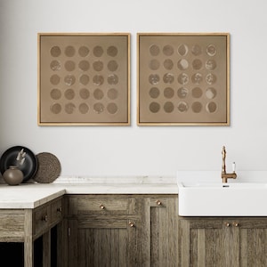 May include: Two framed wall art pieces with a neutral color palette. Each piece features a grid of circular shapes, depicting phases of the moon. The frames are a light brown color, and the background is a muted tan.