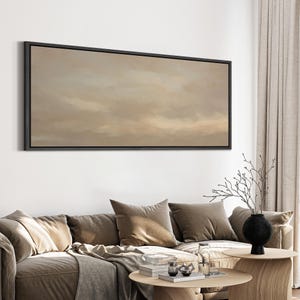 Puede incluir: Una obra de arte enmarcada que representa un paisaje abstracto con un cielo nublado en tonos beige y marr&oacute;n. La obra de arte se exhibe sobre un sof&aacute; con cojines marrones y una mesa de centro de madera.