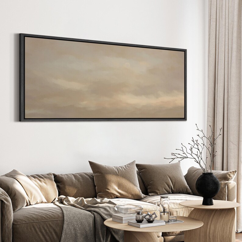 Puede incluir: Una obra de arte enmarcada que representa un paisaje abstracto con un cielo nublado en tonos beige y marr&oacute;n. La obra de arte se exhibe sobre un sof&aacute; con cojines marrones y una mesa de centro de madera.
