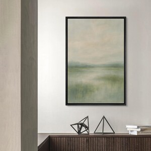 May include: Abstract landscape painting in a black frame. The artwork features a soft, blurred depiction of a natural scene with muted green, beige, and blue tones, evoking a sense of tranquility. The painting is displayed above a wooden cabinet with geometric sculptures.