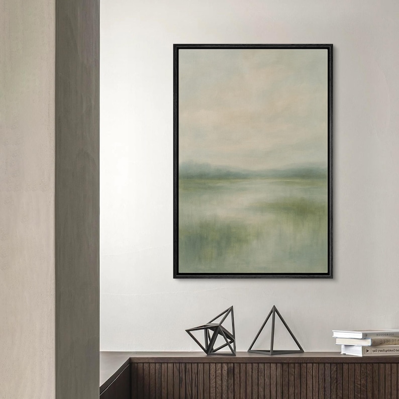 May include: Abstract landscape painting in a black frame. The artwork features a soft, blurred depiction of a natural scene with muted green, beige, and blue tones, evoking a sense of tranquility. The painting is displayed above a wooden cabinet with geometric sculptures.