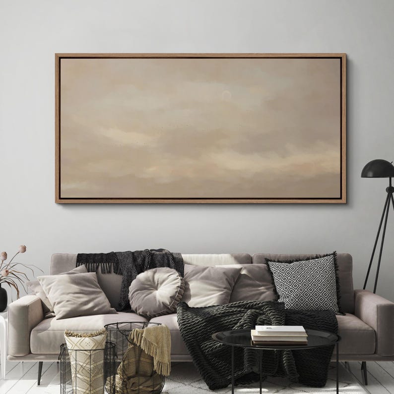 Puede incluir: Una gran obra de arte enmarcada, de forma rectangular, que representa un cielo nublado en tonos beige y marr&oacute;n. La obra de arte cuelga sobre un sof&aacute; con cojines y mantas. La habitaci&oacute;n est&aacute; decorada en tonos neutros.