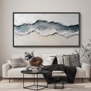 May include: Large abstract wall art with a textured, layered design in shades of white, beige, and blue, framed in black. The artwork hangs above a white sofa with decorative pillows, a black throw, and a black coffee table.