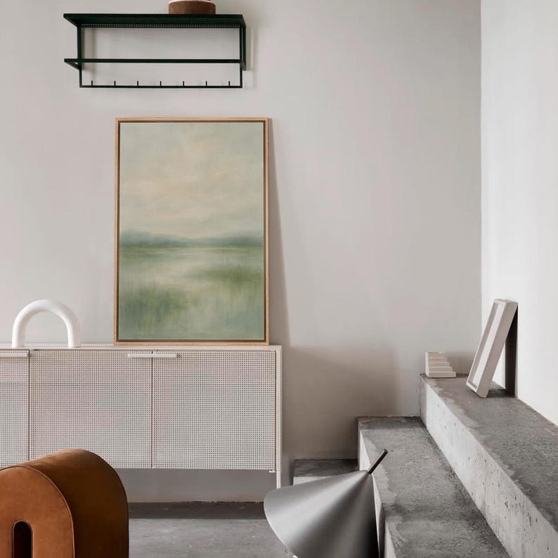 May include: An interior scene featuring a framed landscape painting with a light green and beige color palette. A white cabinet with a perforated design is below the painting. A black shelf and a decorative object are also visible.
