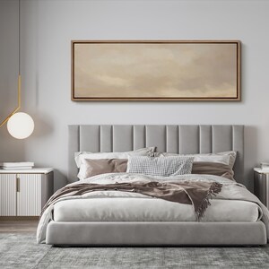 Puede incluir: Un dormitorio moderno con una cama tapizada gris con un cabecero alto con paneles verticales. La cama est&aacute; hecha con ropa de cama blanca y marr&oacute;n, y una manta decorativa. Encima de la cama hay una obra de arte rectangular enmarcada con una paleta de colores neutros.
