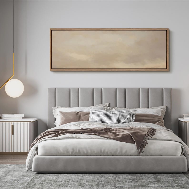 Puede incluir: Un dormitorio moderno con una cama tapizada gris con un cabecero alto con paneles verticales. La cama est&aacute; hecha con ropa de cama blanca y marr&oacute;n, y una manta decorativa. Encima de la cama hay una obra de arte rectangular enmarcada con una paleta de colores neutros.