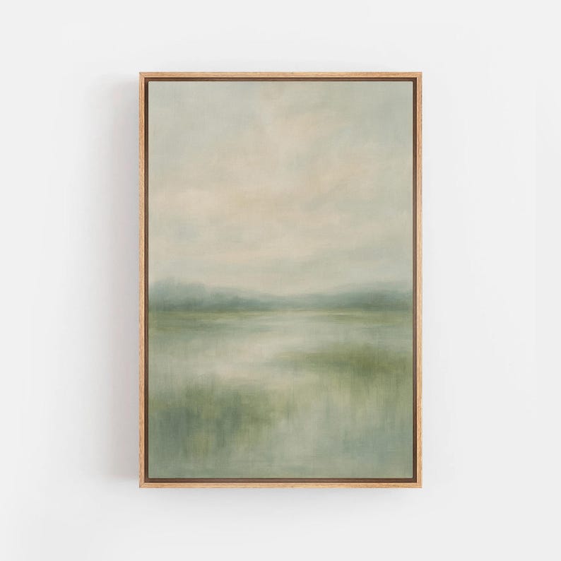 May include: A framed landscape painting with a serene, muted color palette. The artwork depicts a misty scene with soft greens, blues, and beige tones, evoking a sense of calm. The painting is encased in a natural wood frame.
