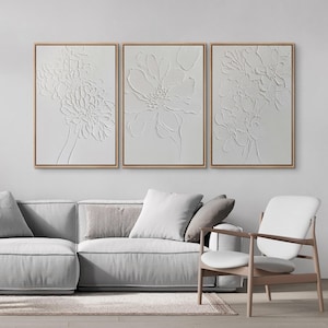 May include: A set of three rectangular wall art pieces featuring white raised floral relief designs on a white background, each enclosed in a thin light wood frame. The textured artwork displays detailed botanical patterns in a monochromatic style.