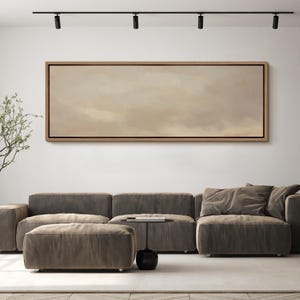 Puede incluir: Una pintura abstracta rectangular grande en un marco de madera cuelga sobre un moderno sof&aacute; seccional gris. La obra de arte presenta una paleta de colores apagados de tonos beige y marr&oacute;n. Una peque&ntilde;a mesa auxiliar negra y una otomana completan el sal&oacute;n.