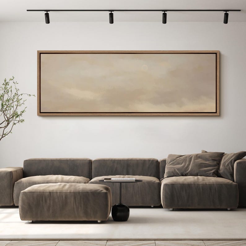 Puede incluir: Una pintura abstracta rectangular grande en un marco de madera cuelga sobre un moderno sof&aacute; seccional gris. La obra de arte presenta una paleta de colores apagados de tonos beige y marr&oacute;n. Una peque&ntilde;a mesa auxiliar negra y una otomana completan el sal&oacute;n.