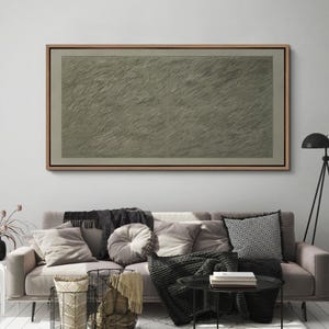 May include: A large, rectangular abstract artwork in a wooden frame. The artwork features a textured, gray-green surface with diagonal lines. The piece is displayed above a sofa with various pillows and blankets.