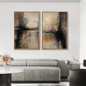 May include: Two framed abstract paintings with a neutral color palette of beige, black, and brown. The artwork features textured brushstrokes and a reflective quality, creating a modern and sophisticated aesthetic. The paintings are displayed above a gray sofa.