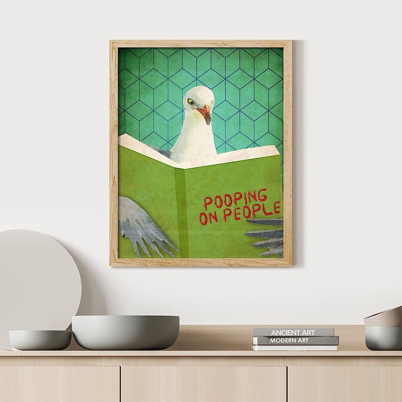 K&ouml;nnte beinhalten: Ein gerahmter Druck einer M&ouml;we, die ein Buch mit dem Titel "POOPING ON PEOPLE" in roten Buchstaben liest. Das Buch ist gr&uuml;n und der Hintergrund ist ein t&uuml;rkisfarbenes geometrisches Muster.