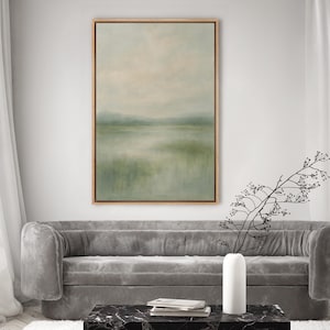 May include: An abstract landscape painting in a wooden frame. The artwork features a serene scene with a muted color palette of greens, blues, and beige, depicting a body of water and distant hills under a hazy sky. The painting is displayed above a gray sofa.