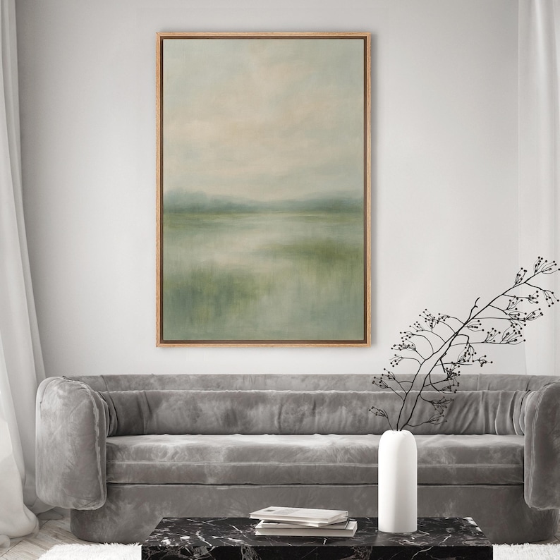 May include: An abstract landscape painting in a wooden frame. The artwork features a serene scene with a muted color palette of greens, blues, and beige, depicting a body of water and distant hills under a hazy sky. The painting is displayed above a gray sofa.