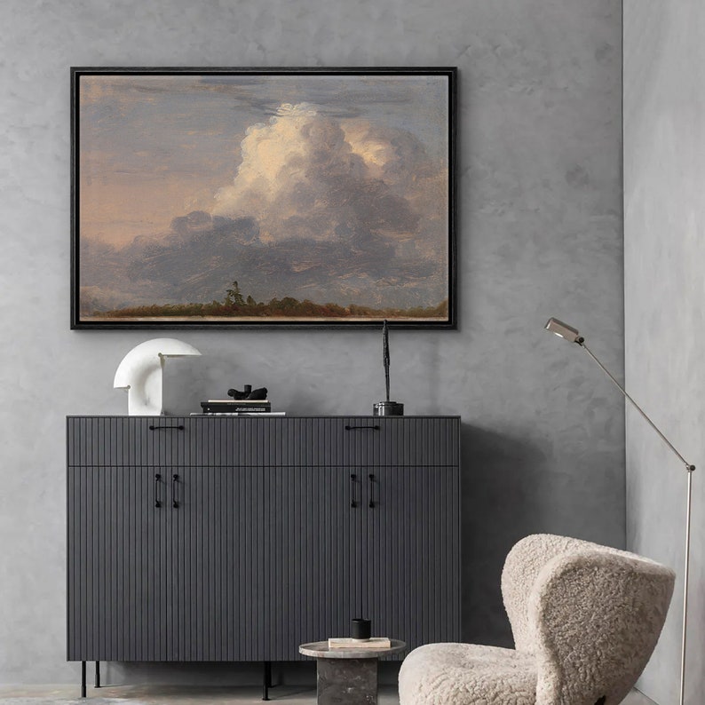 May include: A framed painting of a landscape with a large, fluffy white cloud in the sky. The painting is hanging on a gray wall above a dark gray cabinet with two doors. There is a white lamp on the cabinet and a white chair in the foreground.