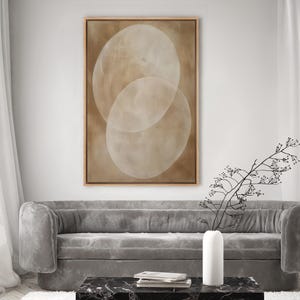 May include: Abstract art print featuring two overlapping, oval shapes in shades of beige and cream. The artwork is framed in a light brown wooden frame and hangs above a gray sofa. The overall aesthetic is minimalist and modern.