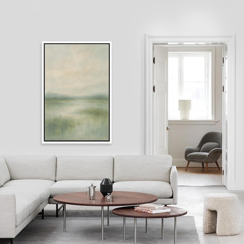 May include: A framed landscape painting with a white border. The artwork depicts a serene scene with soft green and beige hues, suggesting a misty lake or marshland. The painting hangs above a modern sectional sofa.