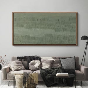 May include: Abstract landscape painting in a wooden frame. The artwork features a muted green color palette with vertical and horizontal brushstrokes, creating a textured effect. The painting is displayed above a sofa with various pillows and blankets, enhancing the room's decor.