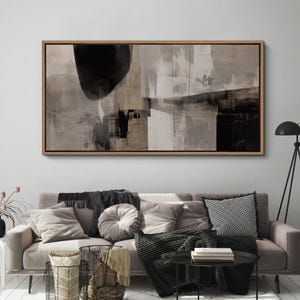 May include: Abstract art print in a wooden frame. The artwork features a composition of black, white, and beige shapes. The painting has a modern, minimalist aesthetic, suitable for home decor. The rectangular canvas is displayed above a sofa.